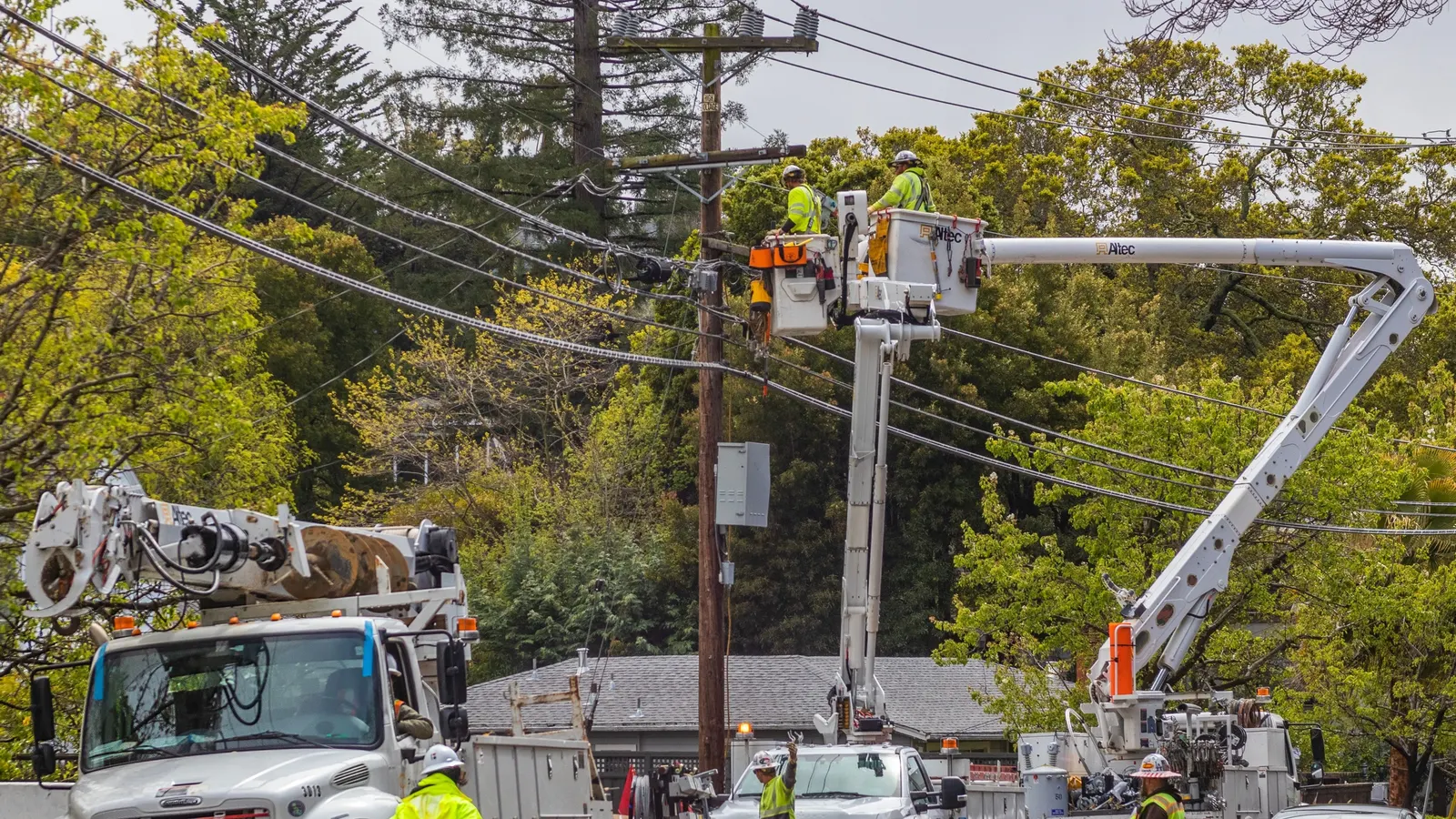 Two linemen in a bucket truck working at a wooden utility pole, with more trucks and traffic cones on the street below